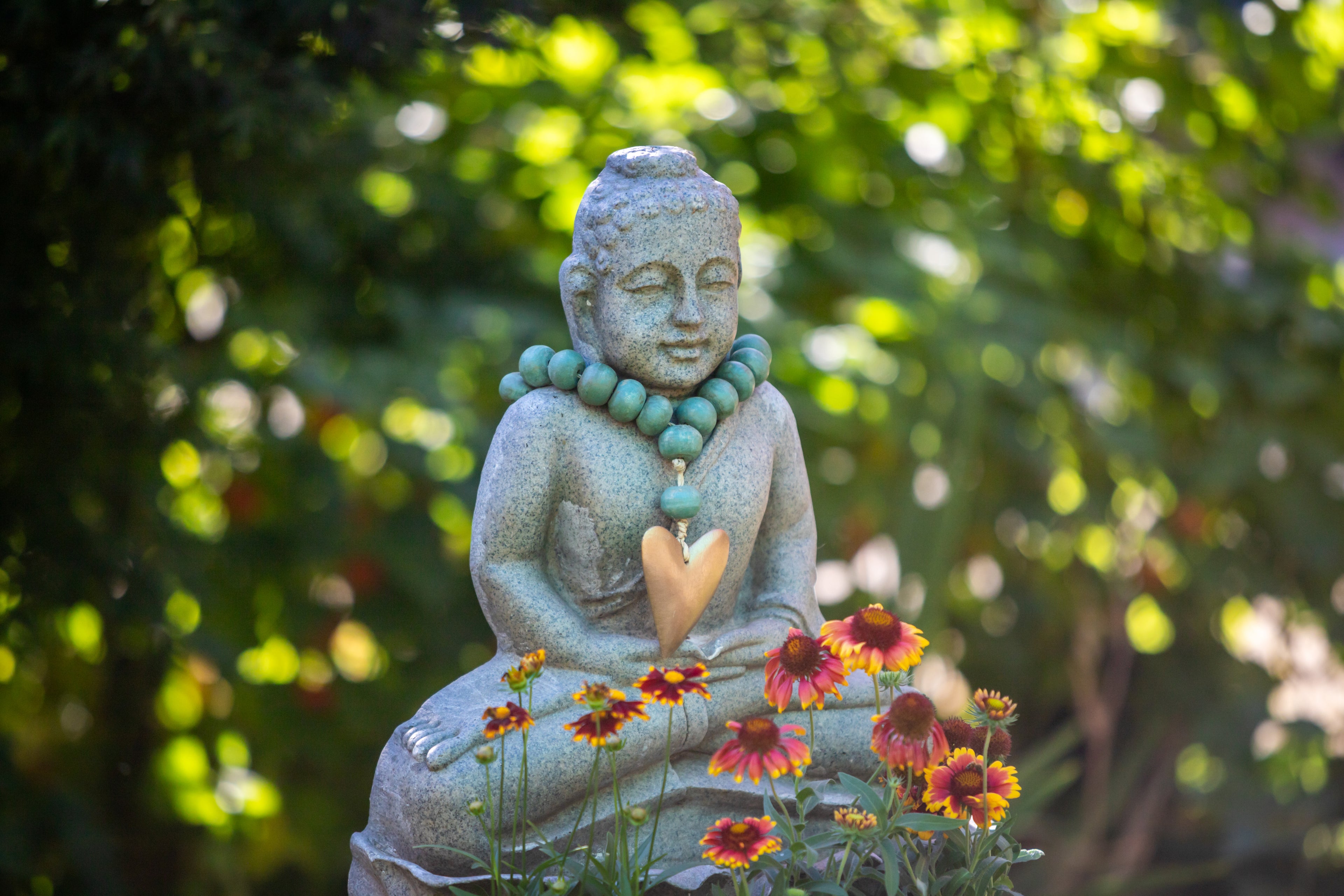 A small statue of a monk meditating sitting behind red and yellow flowers wearing a gold heart necklace. 